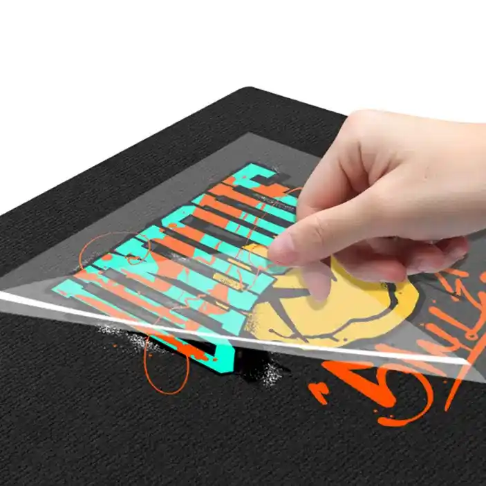 A hand peeling a clear protective plastic film off a freshly heat-pressed Direct-to-Film (DTF) transfer print on a black fabric surface, revealing vibrant street-art graphics.
