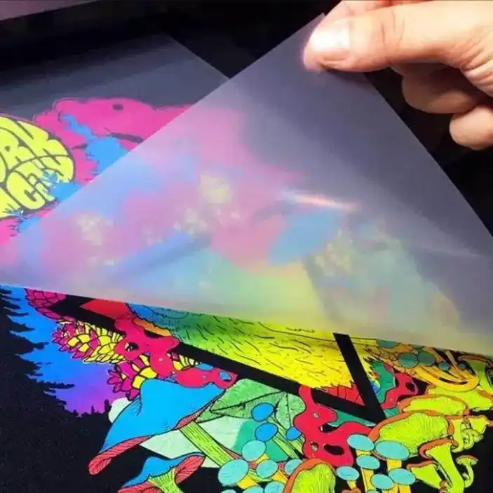 A hand peeling a clear protective plastic film off a freshly heat-pressed Direct-to-Film (DTF) transfer print on a black fabric surface, revealing vibrant street-art graphics.